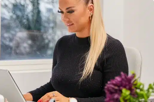 A woman with long blonde hair tied in a ponytail sits at a desk, smiling while working on a laptop. She is wearing a black sweater and there are purple flowers on the desk beside her.