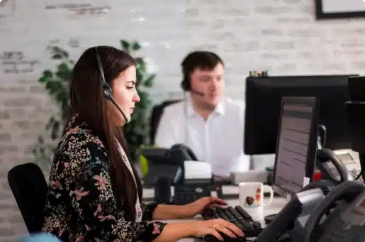 Two office workers wearing headsets sit at desks with computers and phones, focusing on their screens. One woman is in the foreground, typing, while a man is blurred in the background.